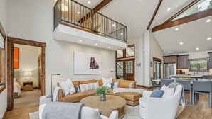 A spacious living area with a large sectional sofa, a coffee table, and a dining area with modern chairs. A staircase leads to an upper level with a balcony.