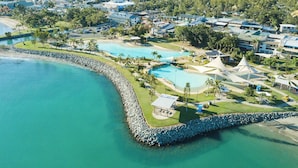 An aerial view of a coastal resort with multiple swimming pools, a rocky shoreline, and a backdrop of residential buildings. - Airlie Sun & Sand #6 (Shute Harbour)