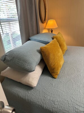 En seng med et quiltet dækken, to puder, en sengetavlelampe og et vindue med persienner.