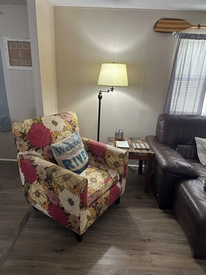 En stue med en blomstret lænestol, en mørk lædersofa, en lampe og et skilt på væggen.