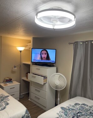 A hotel room with a bed, a desk with a lamp, a TV on a dresser, a fan, and a ceiling light.