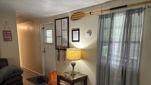 A room with a wooden floor, a brown couch, a small table with a lamp, a door with a bulletin board, and a window with grey curtains. - Rainbow River Experience. (Dunnellon)