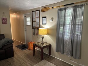 Ein Zimmer mit Holzboden, einem braunen Sofa, einem kleinen Tisch mit Lampe, einer Tür mit Korkbrett und einem Fenster mit grauen Vorhängen.
