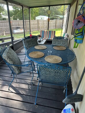 A covered outdoor patio with a round table and chairs, a wooden deck, and a view of a backyard.