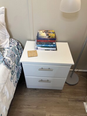 Ein Nachttisch mit Büchern und einer Holzschale, eine Nachttischlampe und ein Bett mit einem gemusterten Überwurf.