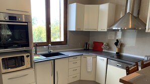 A compact kitchen with a Bosch oven, a microwave, a sink, and a dishwasher. There is a window providing natural light, and a red cutting board is visible on the countertop.