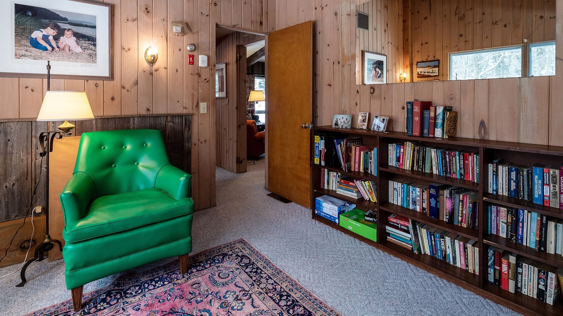 A room with a green leather armchair, a bookshelf filled with books, and a wooden wall with framed pictures.