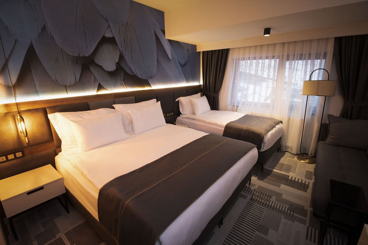 Ein Hotelzimmer mit zwei Betten, einem großen, abstrakten Wandbild darüber, einer Stehlampe und Blick ins Freie durch transparente Vorhänge.