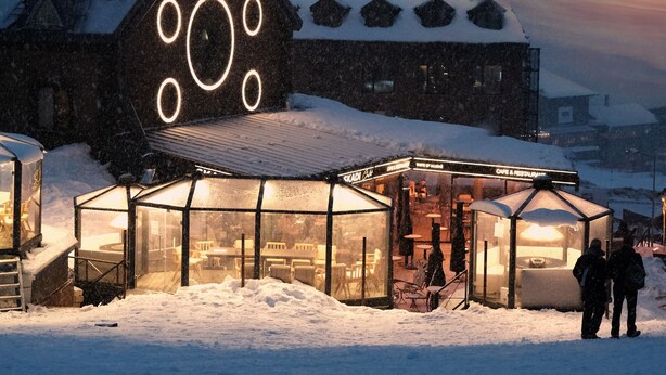 Een gebouw bedekt met sneeuw, met een opvallend raamontwerp in cirkelvorm, een constructie met glazen dak en een bord, en een besneeuwd landschap in de schemering.