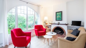 A living room with a fireplace, red chairs, a beige sofa, and a large window overlooking greenery.