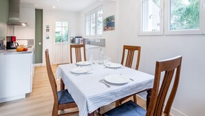 A dining area with a table set for four, wooden chairs, a kitchen in the background, and a view of greenery outside.