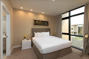 A hotel room with a large bed, a nightstand with a lamp, a framed picture on the wall, and a view of the street through a large window.
