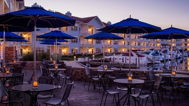 An outdoor dining area with tables and chairs, blue umbrellas, and a marina with boats in the background. - Vintage, restored home w/ pool, hot tub & courtyard-just blocks to town & beach (New Buffalo)