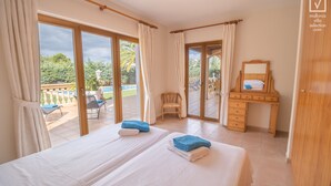Ein Hotelzimmer mit zwei Betten, einem Holztisch, einem Stuhl und einem Balkon mit Blick auf Grünflächen und einen Pool.
