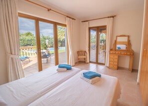 A hotel room with two beds, a wooden desk, a chair, and a balcony with a view of greenery and a pool.