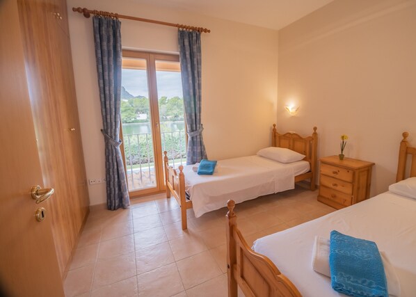A hotel room with two single beds, a wooden wardrobe, a nightstand with a lamp, and a balcony with curtains.