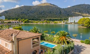 A hotel with a swimming pool, surrounded by greenery and a lake, with mountains in the background.