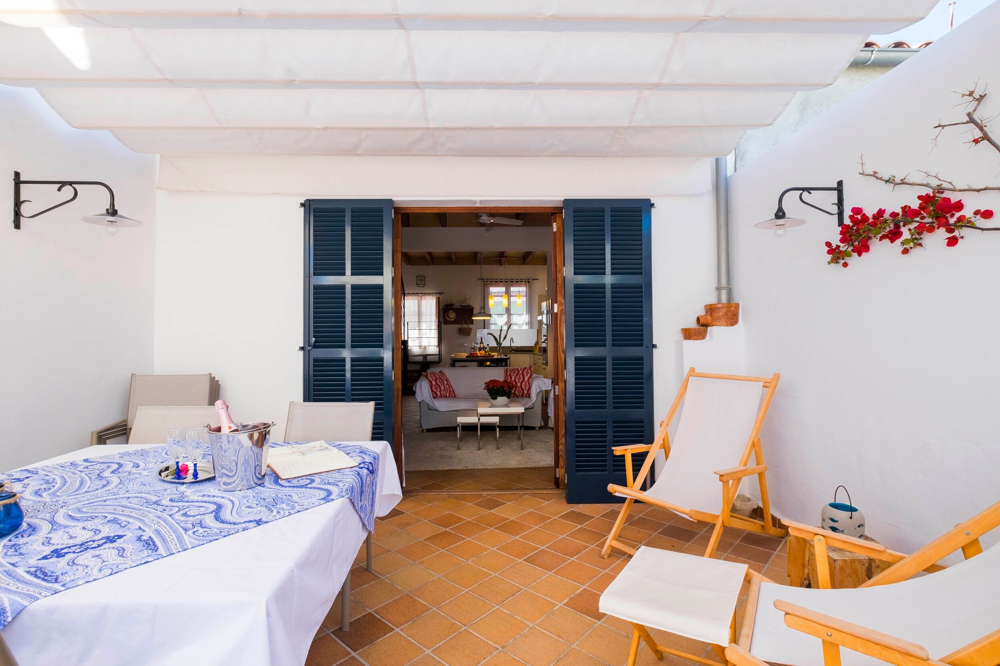 A room with a bed, a table with a tablecloth, a chair, and a view into another room with a sofa and a table.