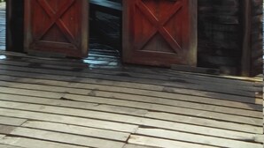 A rustic wooden structure with two large doors, a stone wall, and a roof emitting smoke.