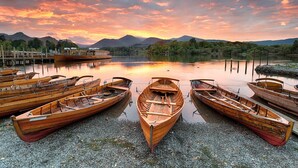 A lakeside scene with wooden boats docked on pebbled shore, a wooden pier, and mountains in the background.