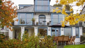 A two-story house with a balcony, a patio with wooden furniture, and a garden with autumn foliage.