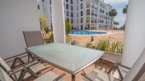 A balcony with a glass table and chairs, overlooking a swimming pool and a multi-story building with balconies. - Marina Real apto 303 (Manilva)