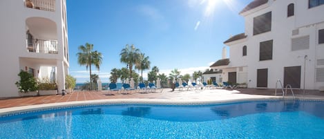 A clear blue pool with lounge chairs and palm trees, surrounded by white buildings with balconies.