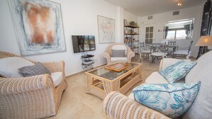 A spacious living room with wicker furniture, a glass coffee table, and artwork on the walls.