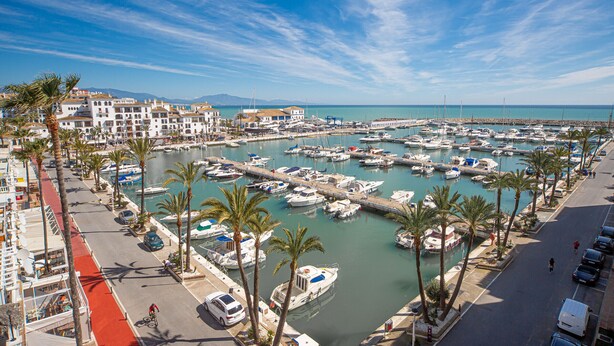 A marina with boats docked, a road alongside, and buildings in the background. - Marina Real number 254 (Manilva)