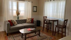 A living room with a sofa, coffee table, and dining table with chairs. There are curtains on the windows, a vase with flowers, and a framed picture on the wall. - Spacious room with private entrance near Bidwell Park! (Chico)