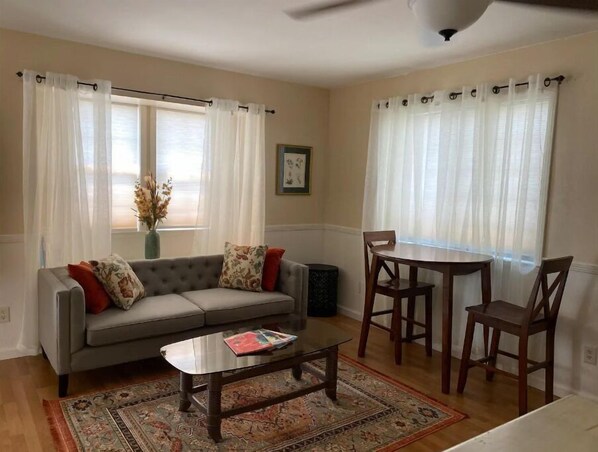 A living room with a sofa, coffee table, and dining table with chairs. There are curtains on the windows, a vase with flowers, and a framed picture on the wall.