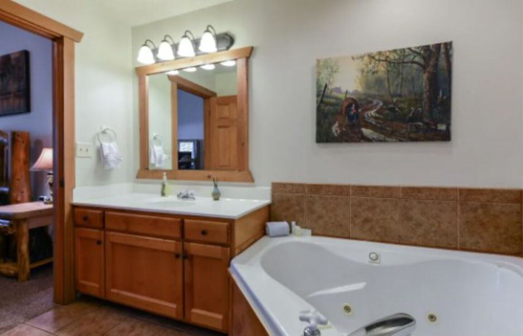 A bathroom with a wooden vanity, a large mirror with lights, a painting of a forest scene, and a white bathtub.
