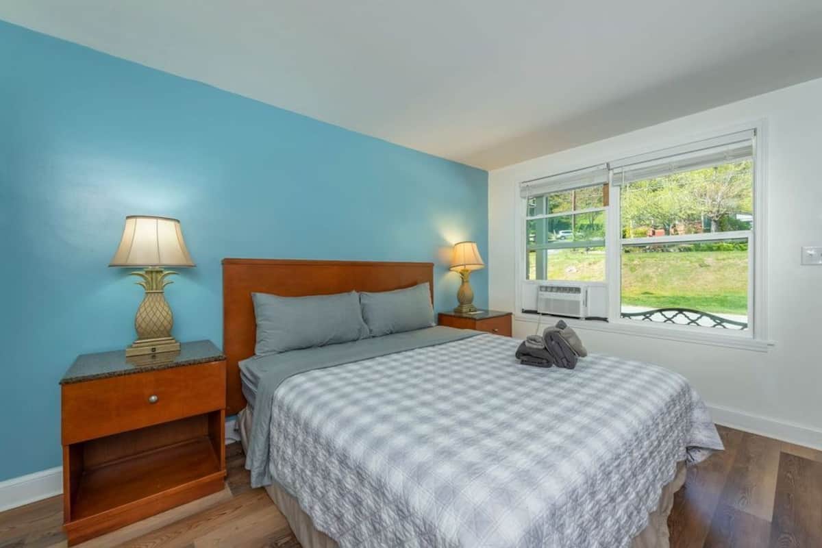 A bedroom with a bed, two bedside tables with lamps, a window with a view of greenery, and a wooden headboard.