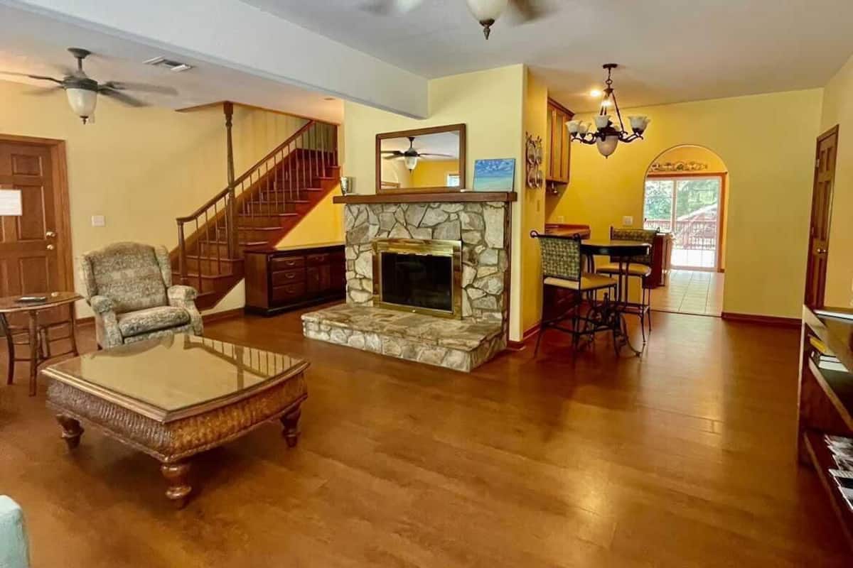 A spacious living room with a stone fireplace, wooden flooring, and a staircase leading to another level.