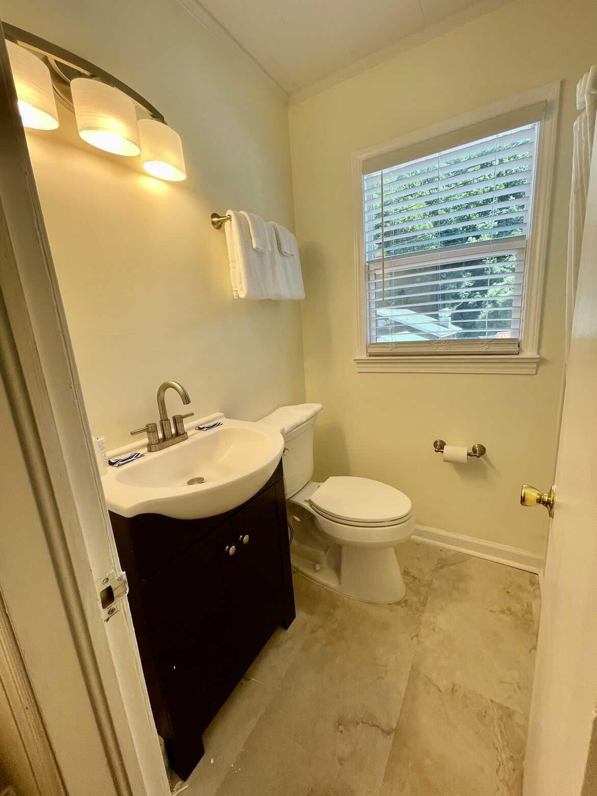 Ein Badezimmer mit weißem Waschbecken, dunklem Waschtisch, Toilette und einem Fenster mit Jalousien.