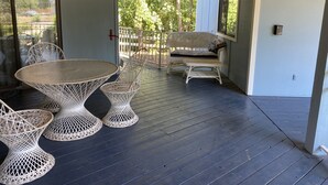 A covered patio with a round table and chairs, a wicker sofa, and a railing overlooking greenery.