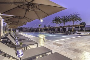 Un espace aménagé au bord de la piscine, avec des chaises longues et des parasols, une piscine et des palmiers.