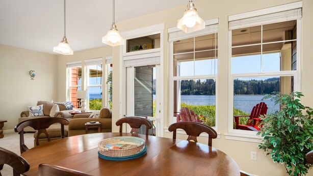 A dining area with a wooden table, chairs, and a view of a lake through large windows. - Happy Place at the Beach (Port Ludlow)