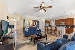 A living room with a sofa, coffee table, and TV, a dining area with chairs, and a kitchen with cabinets and appliances.
