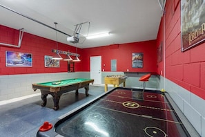 Una mesa de billar, una mesa de fútbol de mesa y una mesa de carambola en una sala con paredes rojas y un proyector.