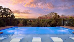 Ein Schwimmbecken mit Liegestühlen, umgeben von Bäumen und Bergen, mit einem Sonnenuntergang im Hintergrund.