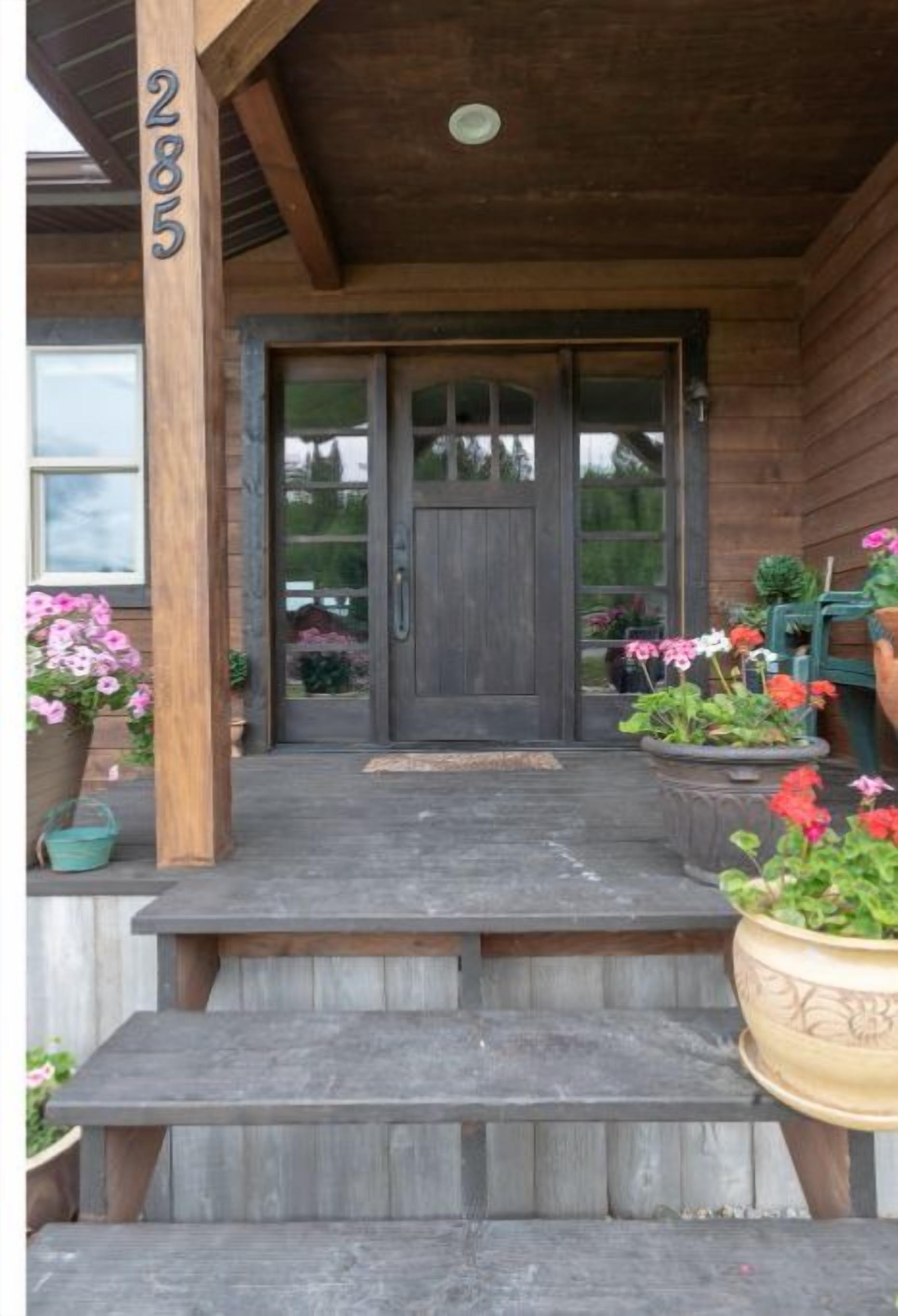 Une véranda en bois à l’avant de la maison, avec une porte sombre, des marches menant à l’entrée et des plantes en pot.