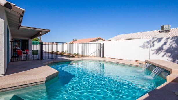 A swimming pool with a clear blue water, a small waterfall feature, and a fenced area with a patio and chairs. - OCO-Heated pool for additional fee and spacious 4br home for your comfort. (Yuma)