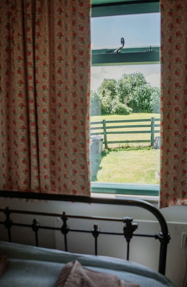 Un dormitorio con vistas a un campo verde y una cerca de madera, visible a través de una ventana con cortinas florales.