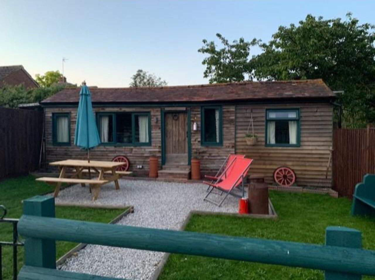 A wooden cabin with a picnic table, a red chair, and a wheelbarrow in the yard.