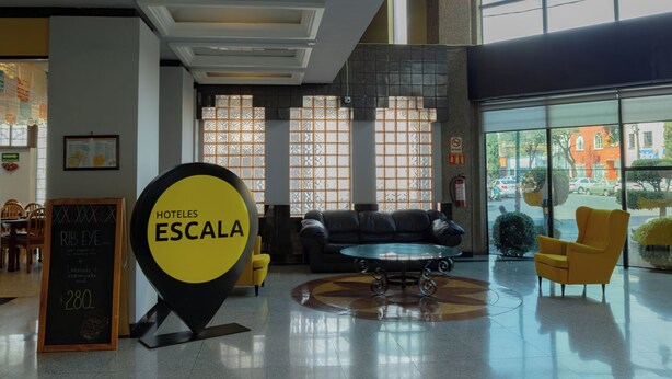 Eine Hotelhalle mit einem großen, gelben Schild, auf dem “HOTELES ESCALA” steht, einer schwarzen Ledersofa, gelben Stühlen und einem Glastisch.