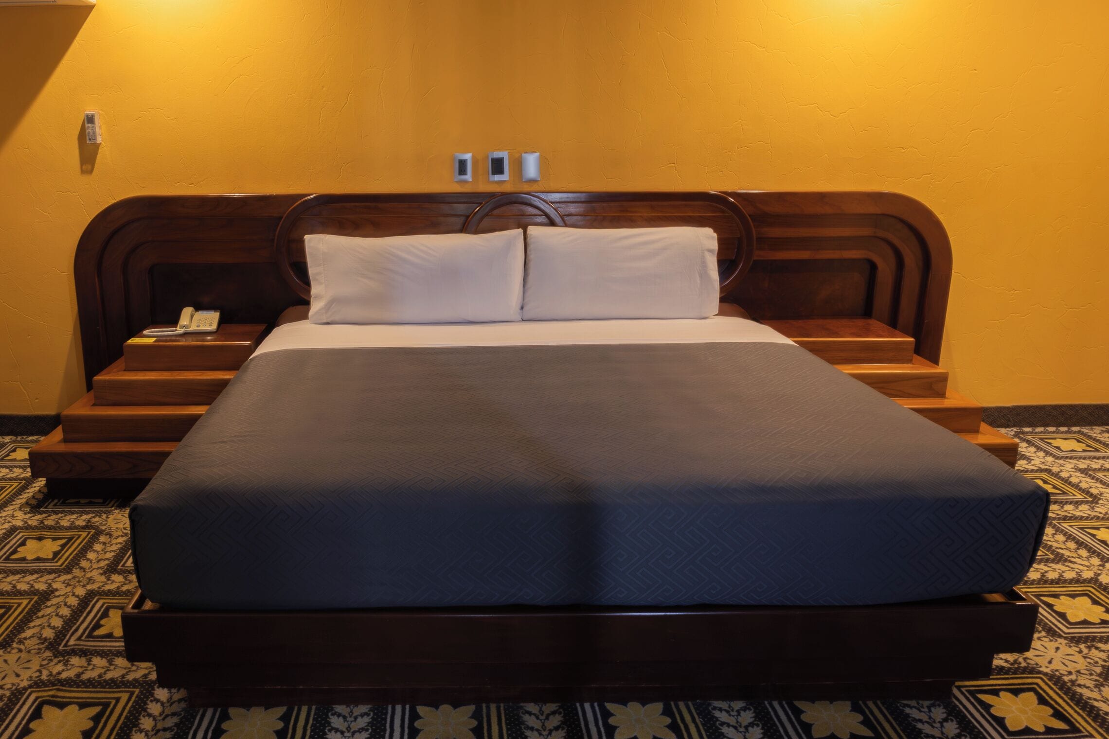 A neatly made bed with a dark blue bedspread and two pillows, a wooden headboard, and a bedside table with a telephone.