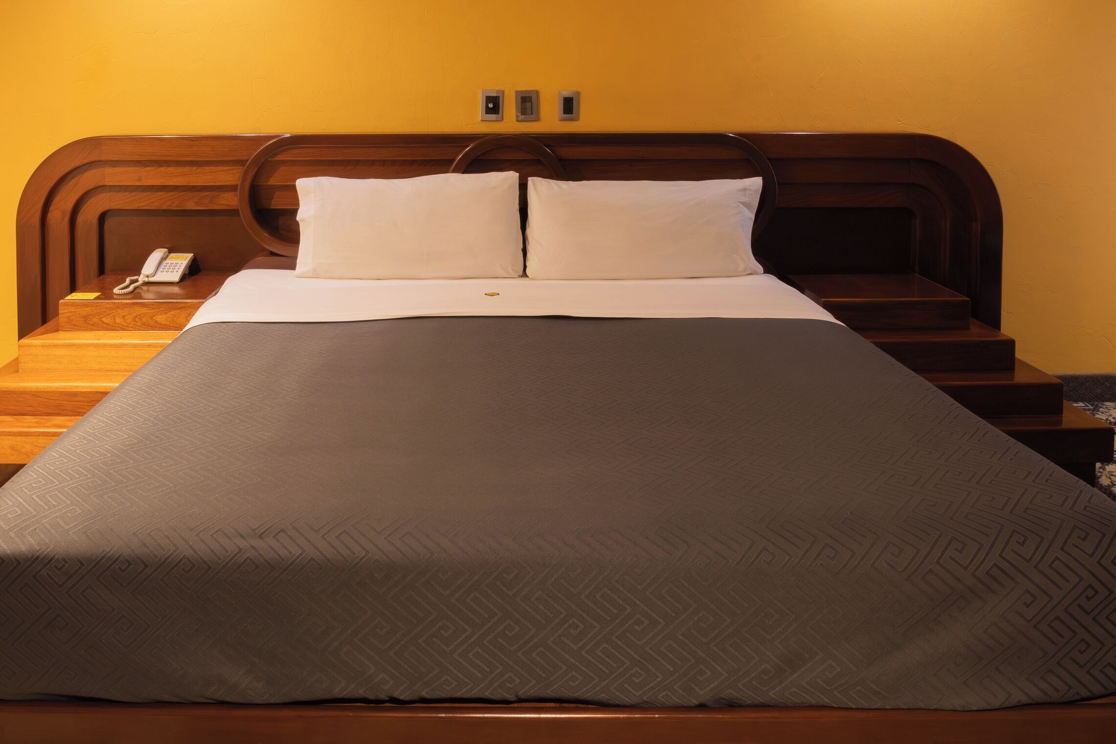 Una cama bien hecha con una colcha estampada, un teléfono en la mesita de noche y un cabecero de madera.