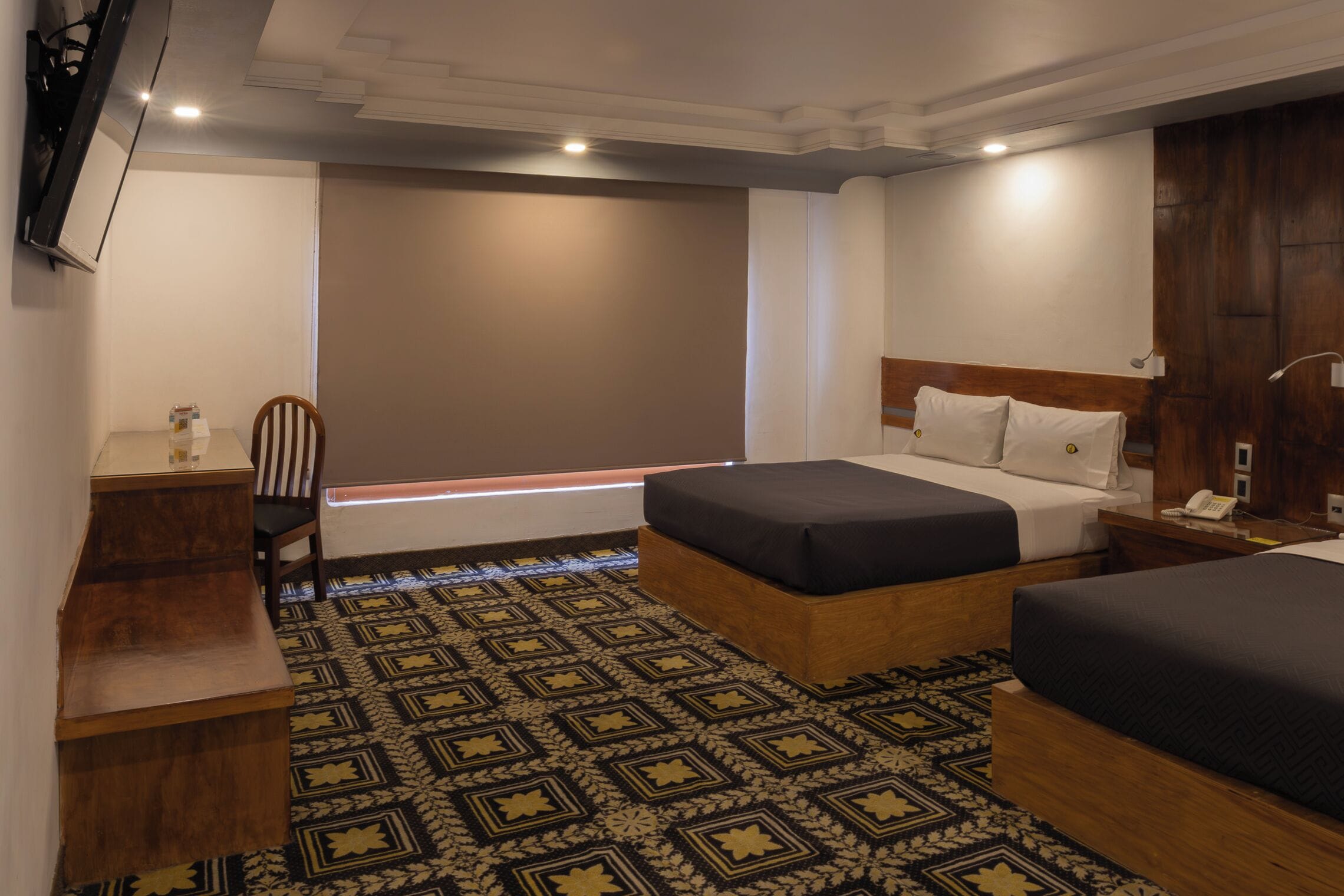 A hotel room with two beds, a desk, a chair, and a patterned carpet.