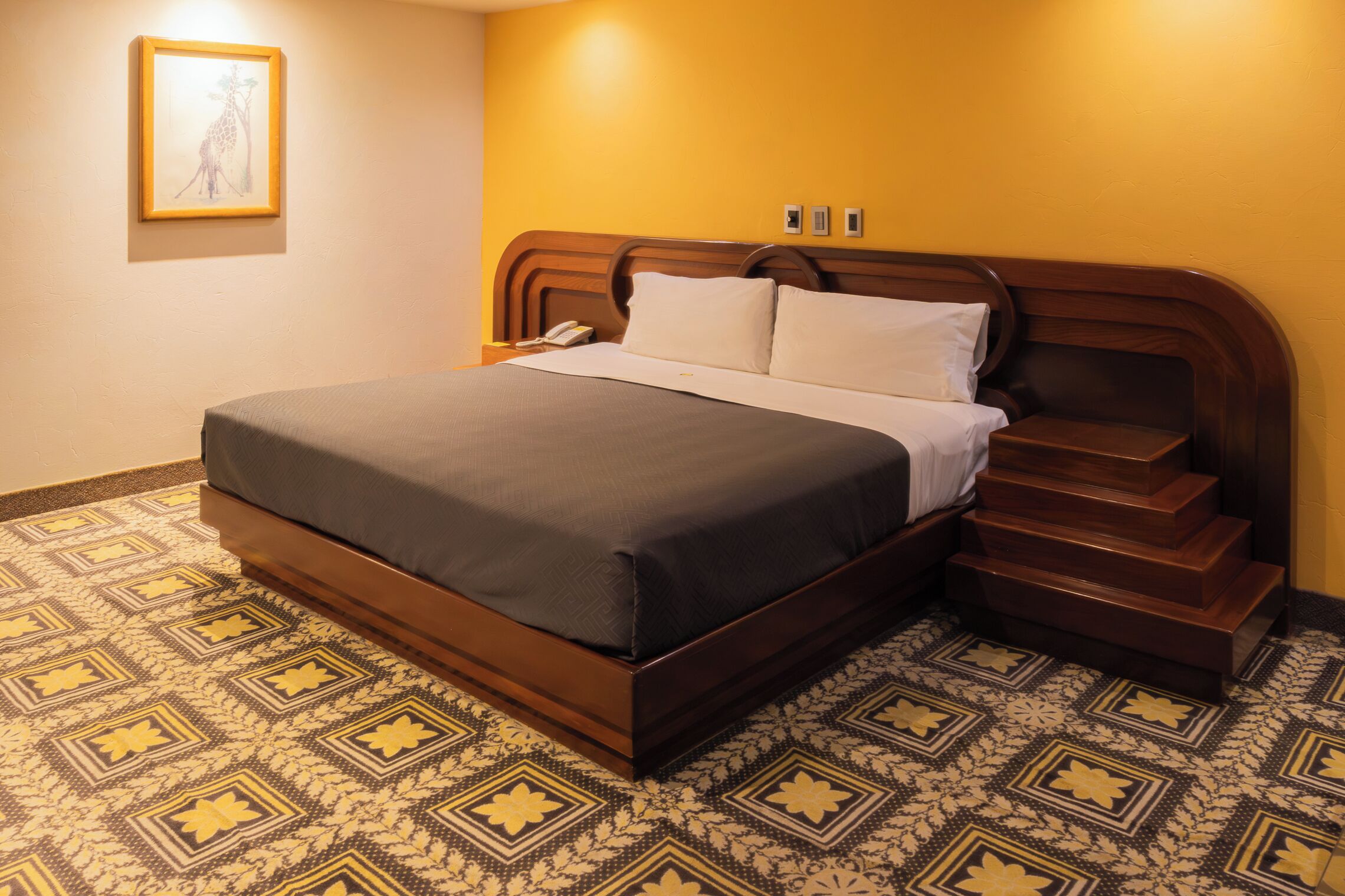 A neatly made bed with a wooden headboard, a patterned floor, and a framed picture on the wall.